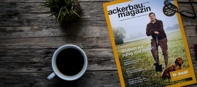 EIne Ausgabe des Ackerbaumagazins liegt auf einem Holztisch neben einer Zimmerpflanze und einer Tasse Kaffee.