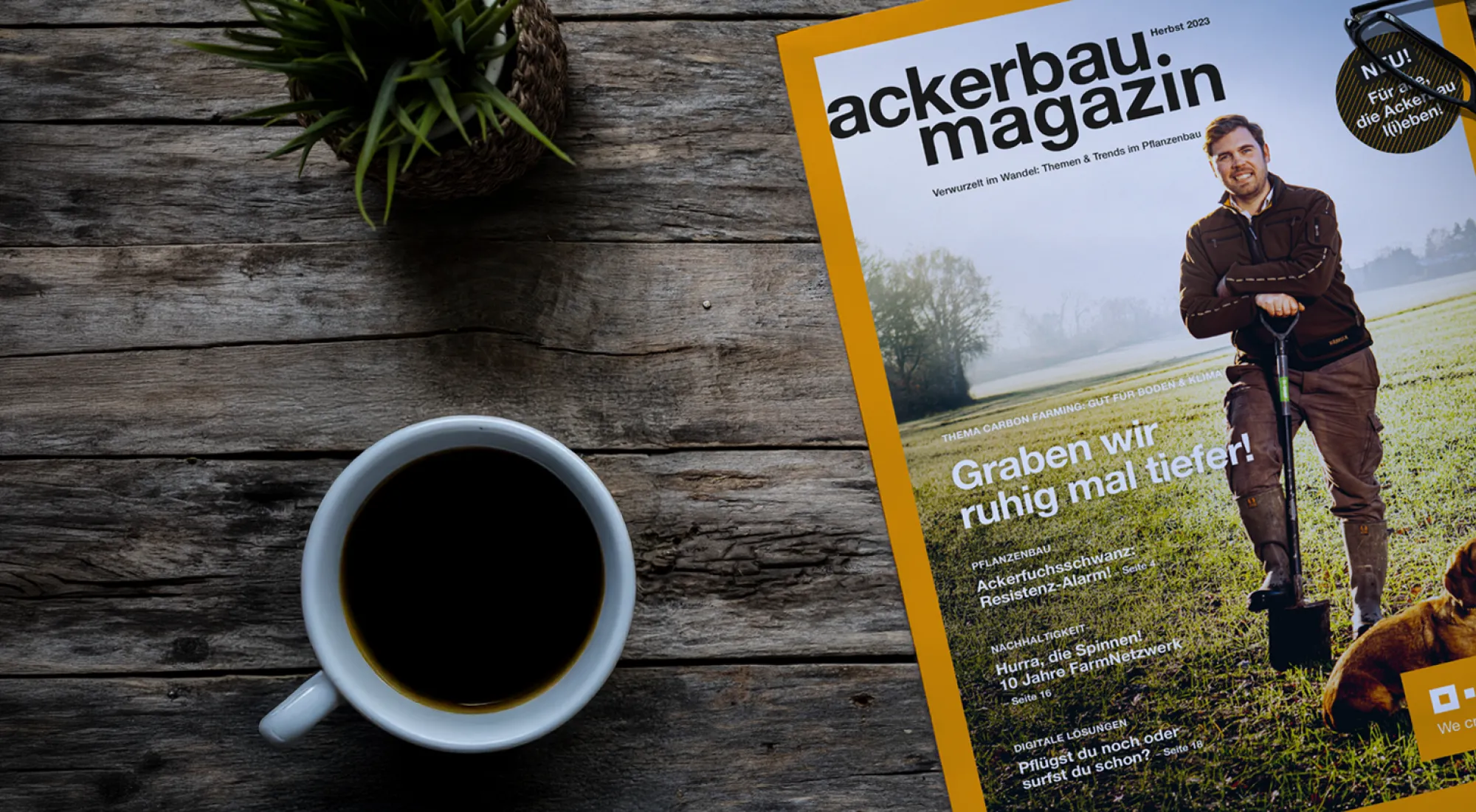 EIne Ausgabe des Ackerbaumagazins liegt auf einem Holztisch neben einer Zimmerpflanze und einer Tasse Kaffee.